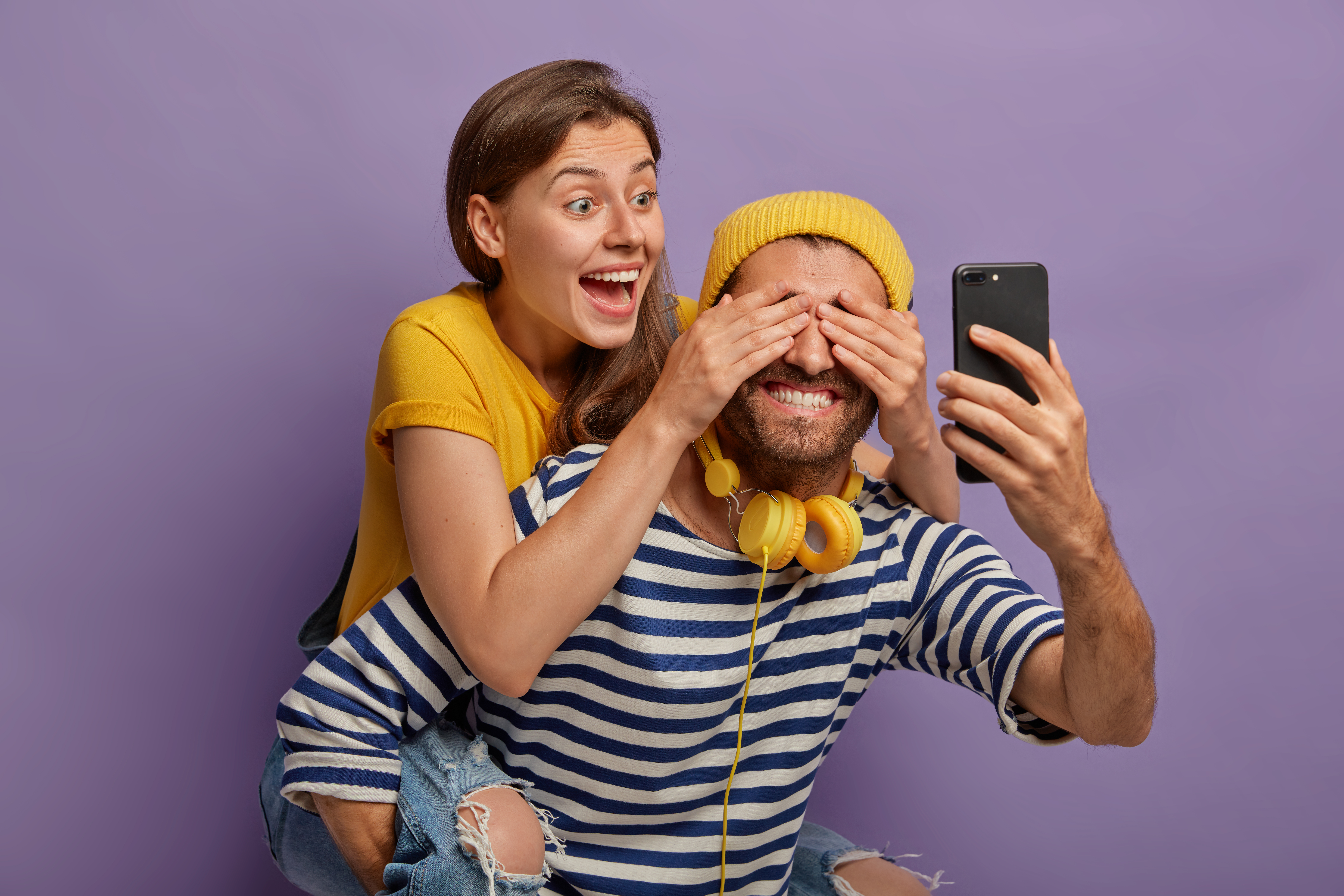 Happy girlfriend rides piggyback, has fun with boyfriend covers his eyes, prepares surprise. Cheerful hipster holds smartphone in front, wears headphones around neck. Couple foolishing around