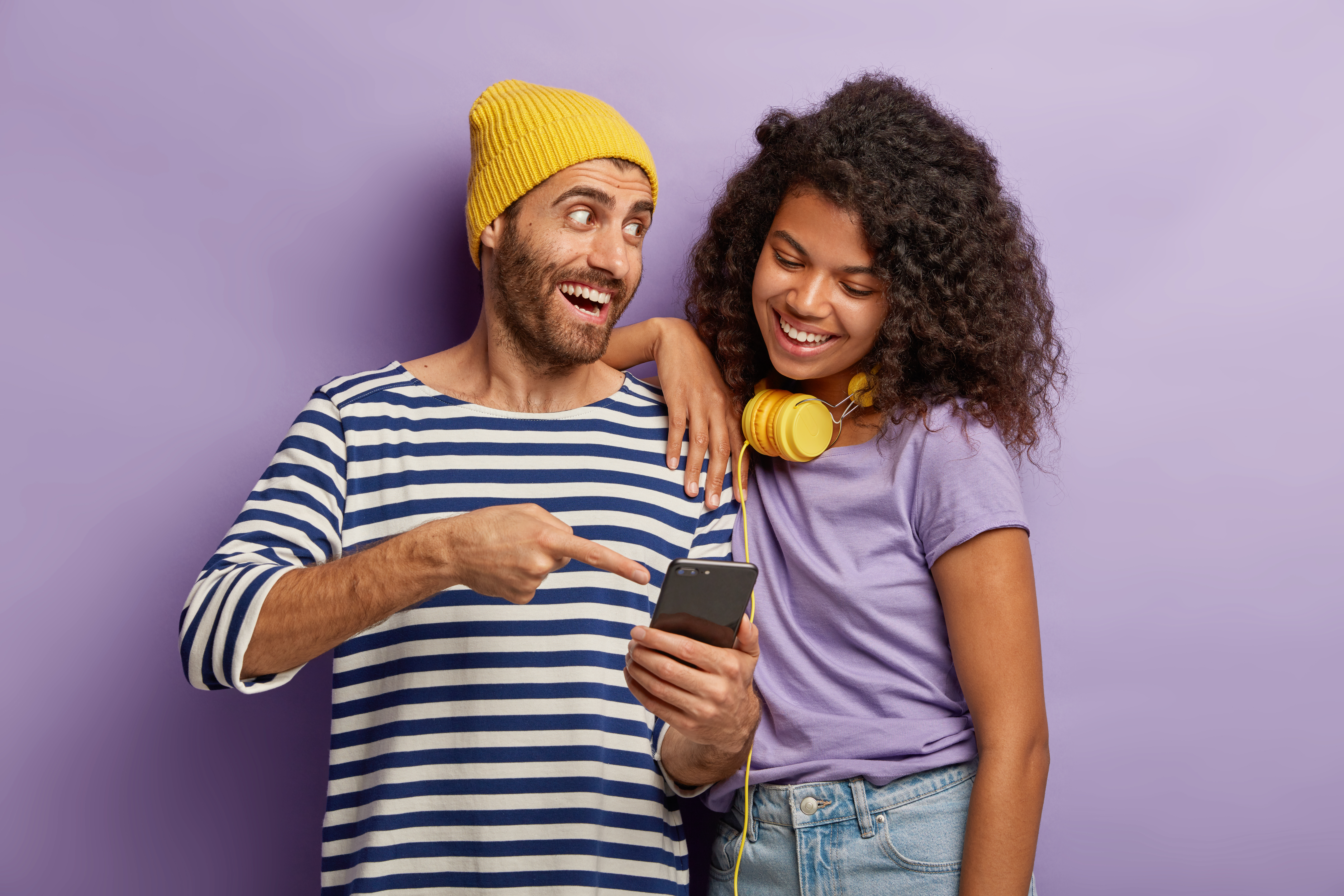 Happy guy in yellow hat and striped jumper, explains to Afro girl how to use new application at smartphone, points in display, stand closely, cannot imagine life without modern technologies.