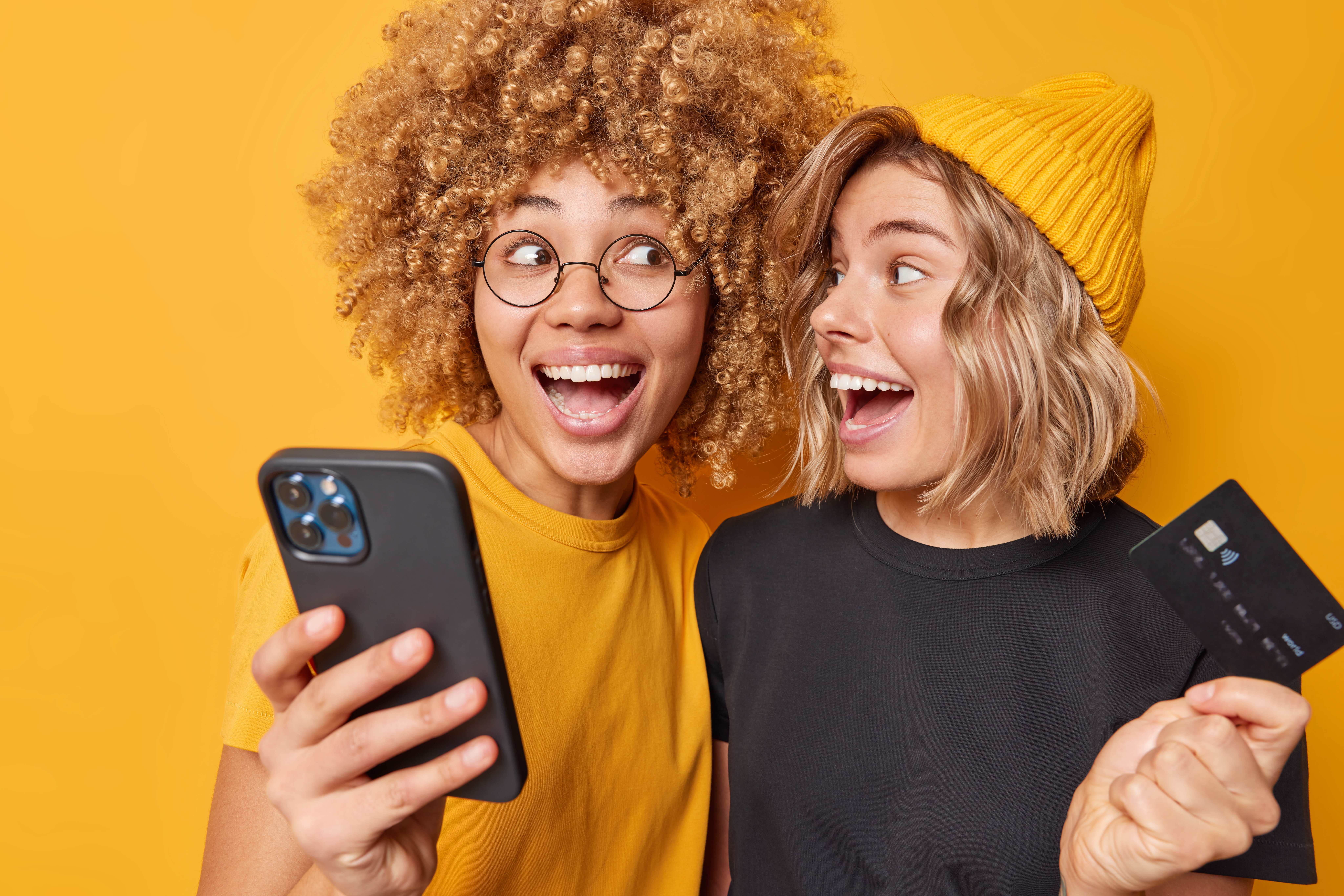 Positive glad female friends holds credit card and smartphone happy to spend money on online shopping purchase goods or services in internet isolated over yellow background check their balance